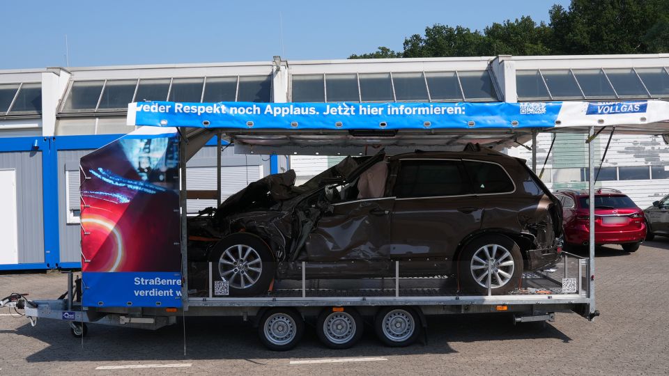 Kampagnenanhänger in Seitenansicht. Die Seitenwände sind hochgeklappt, zu sehen ist ein Fahrzeug mit Totalschaden. Am Dach des Anhängers ist der Schriftzug "Wer Respekt noch Applaus. Jetzt hier informieren" zu sehen