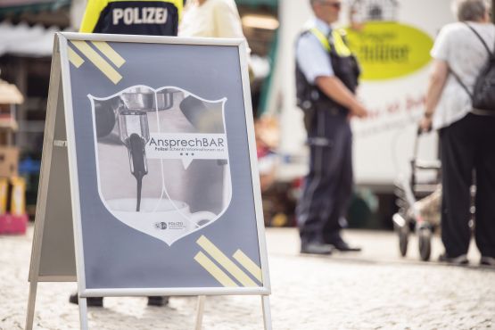 Ein Aufsteller zeigt das Logo der AnsprechBAR - darauf ist eine Tasse Kaffee und der Schriftzug "AnsprechBAR - Polizei schenkt Informationen aus" zu lesen. Im Hintergrund stehen Polizisten und Besucher.